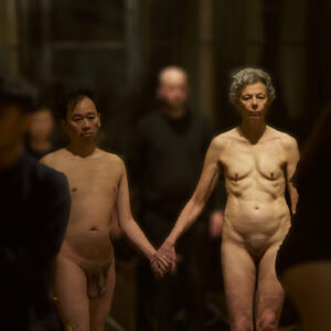 A dramatic art installation scene featuring a man standing nude and a woman kneeling in front of him while an older nude woman stands nearby, all set in a dimly lit room with chandeliers and a blurred audience in the background. Philemon Mukarno Performance Art Co-Creation(2020) 3h Venice International Performance Art Week 2020, Palazzo Mora, Venice, Italia