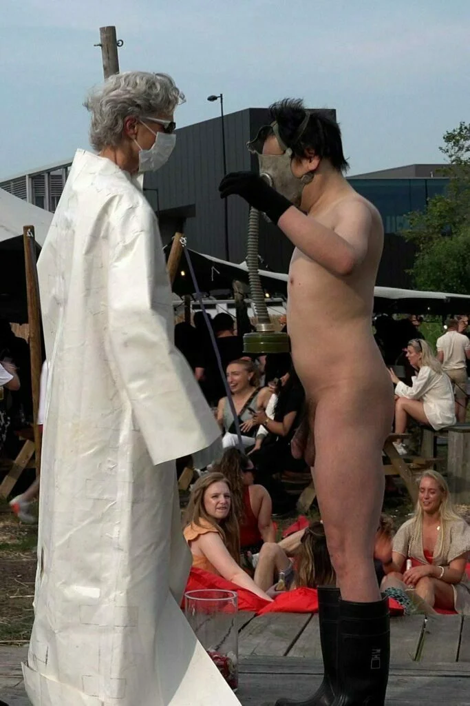 A naked performer wearing a gas mask and black rubber boots embraces another figure dressed in a white outfit, on a wooden platform scattered with flower petals, amidst an outdoor event with spectators in the background. Naked Performance Art De-Coronasation for Spiritual Healing by Ingrid Adriaans and Philemon Mukarno in Amsterdam