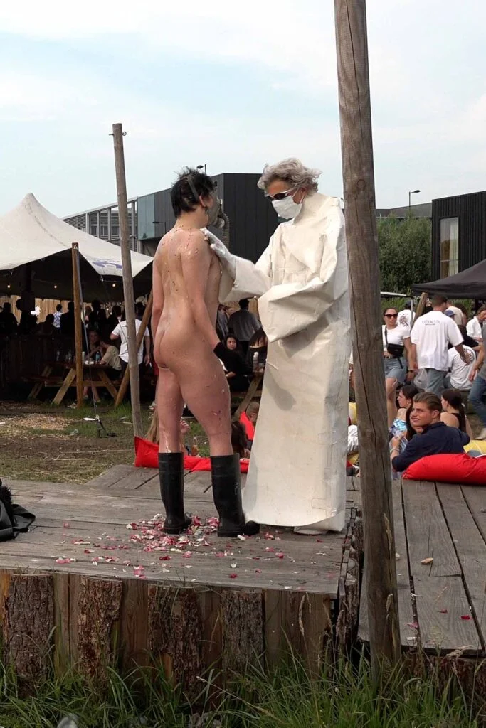 A naked performer wearing a gas mask and black rubber boots embraces another figure dressed in a white outfit, on a wooden platform scattered with flower petals, amidst an outdoor event with spectators in the background. Naked Performance Art De-Coronasation for Spiritual Healing by Ingrid Adriaans and Philemon Mukarno in Amsterdam