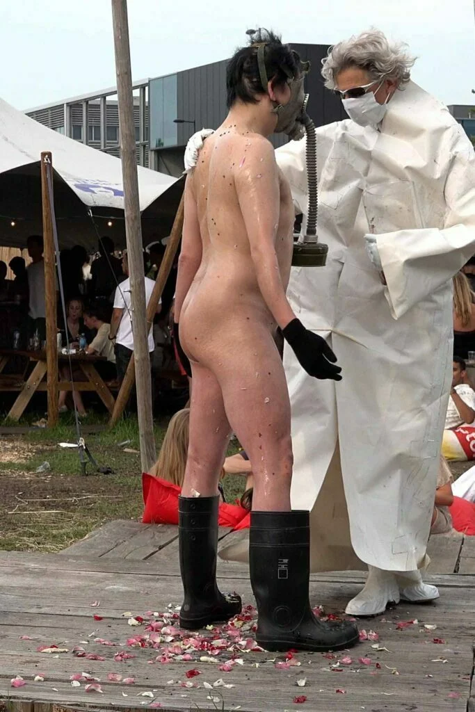 A naked performer wearing a gas mask and black rubber boots embraces another figure dressed in a white outfit, on a wooden platform scattered with flower petals, amidst an outdoor event with spectators in the background. Naked Performance Art De-Coronasation for Spiritual Healing by Ingrid Adriaans and Philemon Mukarno in Amsterdam