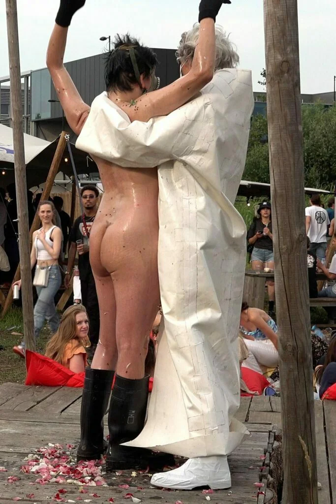 A naked performer wearing a gas mask and black rubber boots embraces another figure dressed in a white outfit, on a wooden platform scattered with flower petals, amidst an outdoor event with spectators in the background. Naked Performance Art De-Coronasation for Spiritual Healing by Ingrid Adriaans and Philemon Mukarno in Amsterdam