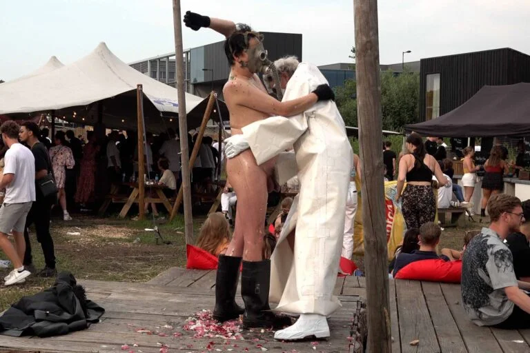 A naked performer wearing a gas mask and black rubber boots embraces another figure dressed in a white outfit, on a wooden platform scattered with flower petals, amidst an outdoor event with spectators in the background. Naked Performance Art De-Coronasation for Spiritual Healing by Ingrid Adriaans and Philemon Mukarno in Amsterdam