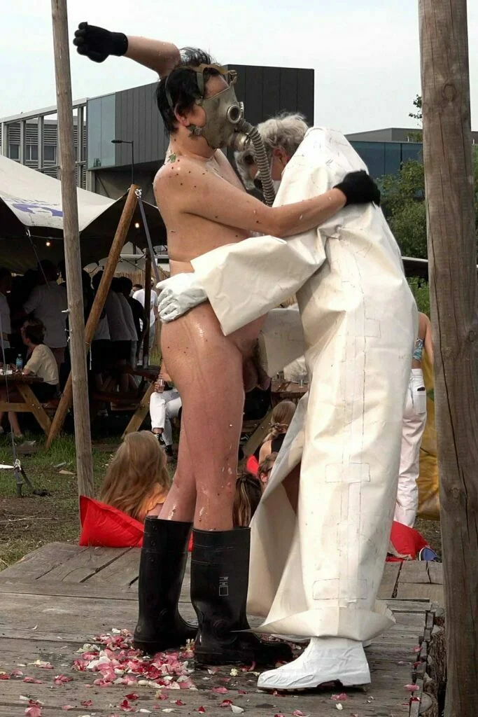 A naked performer wearing a gas mask and black rubber boots embraces another figure dressed in a white outfit, on a wooden platform scattered with flower petals, amidst an outdoor event with spectators in the background. Naked Performance Art De-Coronasation for Spiritual Healing by Ingrid Adriaans and Philemon Mukarno in Amsterdam