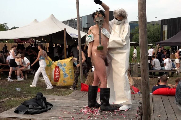 A naked performer wearing a gas mask and black rubber boots embraces another figure dressed in a white outfit, on a wooden platform scattered with flower petals, amidst an outdoor event with spectators in the background. Naked Performance Art De-Coronasation for Spiritual Healing by Ingrid Adriaans and Philemon Mukarno in Amsterdam