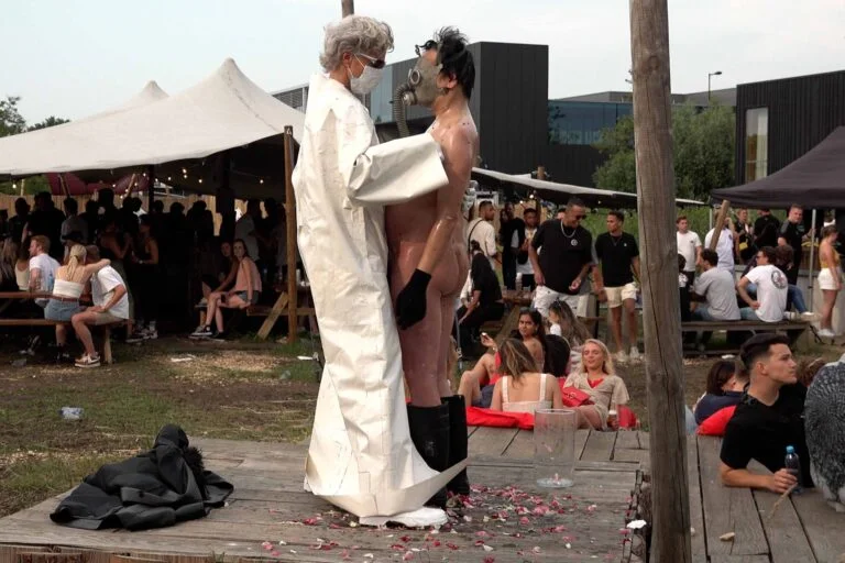 A naked performer wearing a gas mask and black rubber boots embraces another figure dressed in a white outfit, on a wooden platform scattered with flower petals, amidst an outdoor event with spectators in the background. Naked Performance Art De-Coronasation for Spiritual Healing by Ingrid Adriaans and Philemon Mukarno in Amsterdam