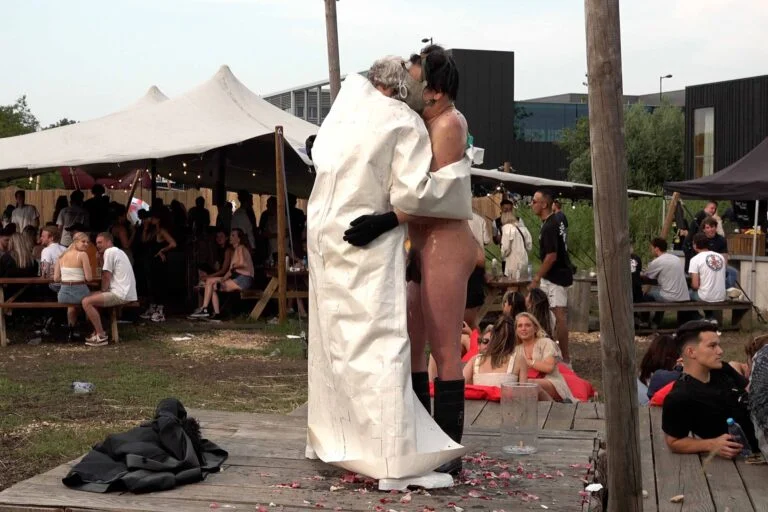 A naked performer wearing a gas mask and black rubber boots embraces another figure dressed in a white outfit, on a wooden platform scattered with flower petals, amidst an outdoor event with spectators in the background. Naked Performance Art De-Coronasation for Spiritual Healing by Ingrid Adriaans and Philemon Mukarno in Amsterdam