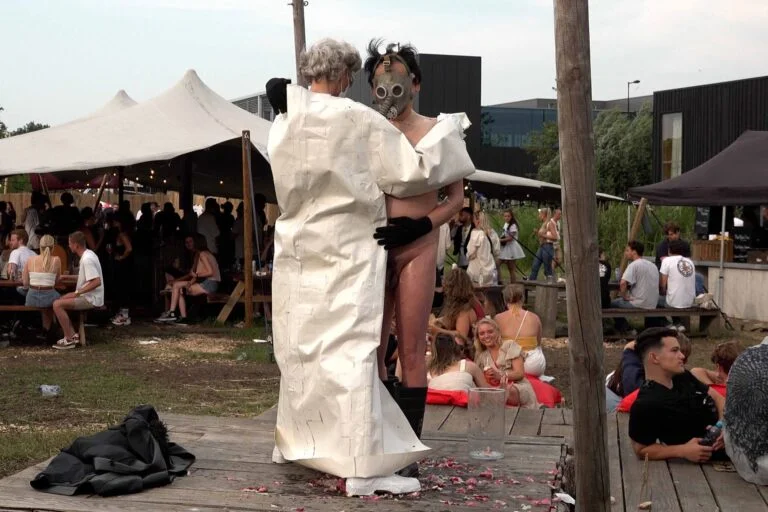 A naked performer wearing a gas mask and black rubber boots embraces another figure dressed in a white outfit, on a wooden platform scattered with flower petals, amidst an outdoor event with spectators in the background. Naked Performance Art De-Coronasation for Spiritual Healing by Ingrid Adriaans and Philemon Mukarno in Amsterdam