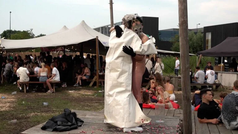 A naked performer wearing a gas mask and black rubber boots embraces another figure dressed in a white outfit, on a wooden platform scattered with flower petals, amidst an outdoor event with spectators in the background. Naked Performance Art De-Coronasation for Spiritual Healing by Ingrid Adriaans and Philemon Mukarno in Amsterdam