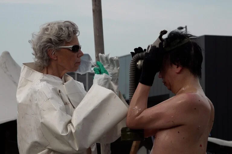 A naked performer wearing a gas mask and black rubber boots embraces another figure dressed in a white outfit, on a wooden platform scattered with flower petals, amidst an outdoor event with spectators in the background. Naked Performance Art De-Coronasation for Spiritual Healing by Ingrid Adriaans and Philemon Mukarno in Amsterdam