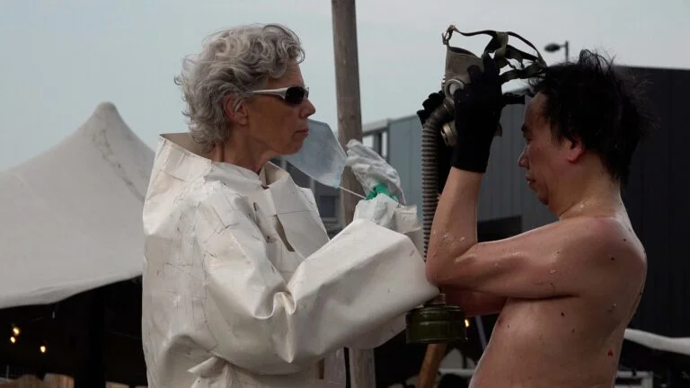 A naked performer wearing a gas mask and black rubber boots embraces another figure dressed in a white outfit, on a wooden platform scattered with flower petals, amidst an outdoor event with spectators in the background. Naked Performance Art De-Coronasation for Spiritual Healing by Ingrid Adriaans and Philemon Mukarno in Amsterdam