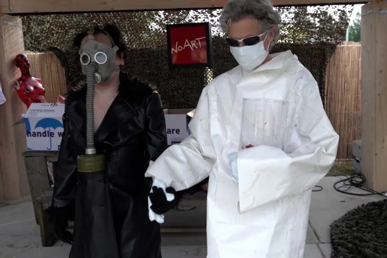 Two individuals dressed in unusual outfits: one wearing a gas mask and a black leather coat, and the other in a white suit with a mask and sunglasses, holding hands. In the background, there is a display with a sign that reads 'NoART' and various items visible. Naked Performance Art De-Coronasation for Spiritual Healing by Ingrid Adriaans and Philemon Mukarno in Amsterdam