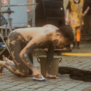 Philemon Mukarno is covered in black paint is on their hands and knees scrubbing a stone pavement with a brush, next to a bucket. A person in the background appears to be wearing a colorful outfit. Transition (2021)'15 Philemon Mukarno Performance.