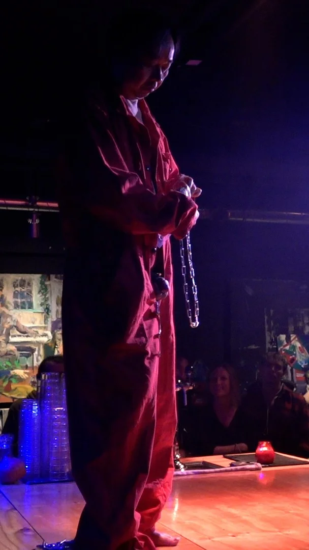 Philemon Mukarno performing Normcore in a red outfit holds a chain and a small object, with an audience visible in the background watching intently, set against a dark backdrop with abstract art. He is on a bar floor, partially covered with a red cloth, is crawling towards a bottle of ketchup on the counter, with colorful ambient lighting and a drink station in the background.