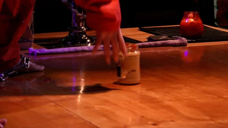 Philemon Mukarno performing Normcore on a bar floor, partially covered with a red cloth, is crawling towards a bottle of ketchup on the counter, with colorful ambient lighting and a drink station in the background. Its a Naked Performance in WORM.