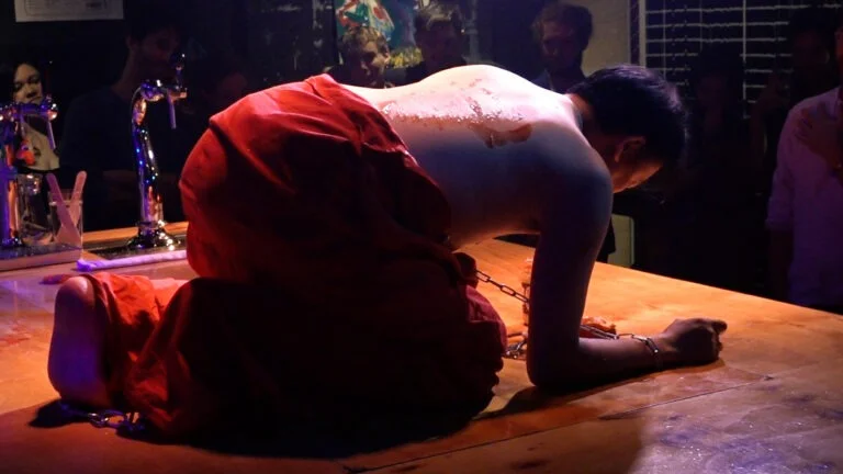 Philemon Mukarno performing Normcore with a bare back, wearing a red garment, kneels on a wooden table chained at the wrists and ankles, with a few visible drops of liquid on their back, while people watch in dim lighting. Its a Naked Performance in WORM.