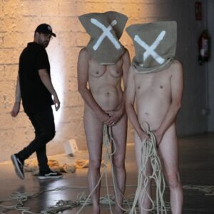 Asphyxia performance art: Two nude figures with burlap sacks over their heads, marked with white 'X's, stand holding lengths of rope in an industrial space. A third person, wearing a black cap and shirt, walks in the background.