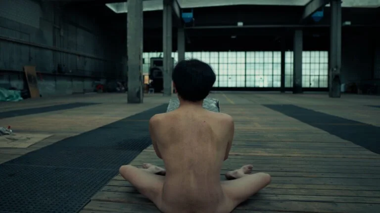 A person sits naked on a wooden floor in a spacious, industrial setting, facing away from the camera and holding a patterned fabric, with large windows in the background letting in natural light.