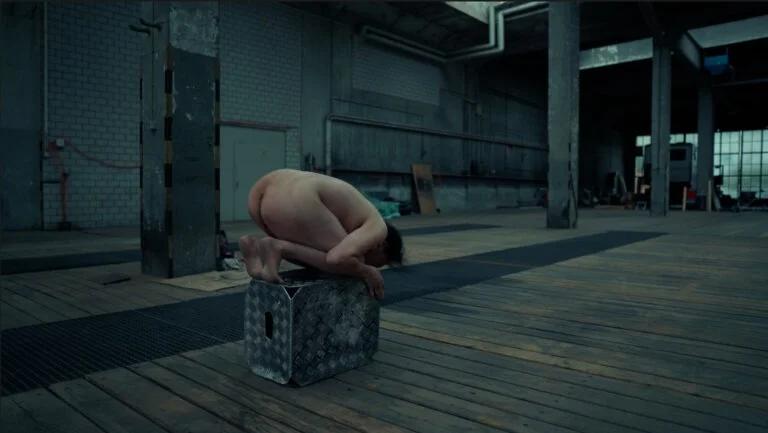 A naked performer is squatting on a stool in a large, empty industrial space with wooden floors and concrete walls. The performer is crouched low, with their head bent down toward their knees, creating a compact shape. The background shows muted lighting and minimal props, contributing to the stark and artistic atmosphere.