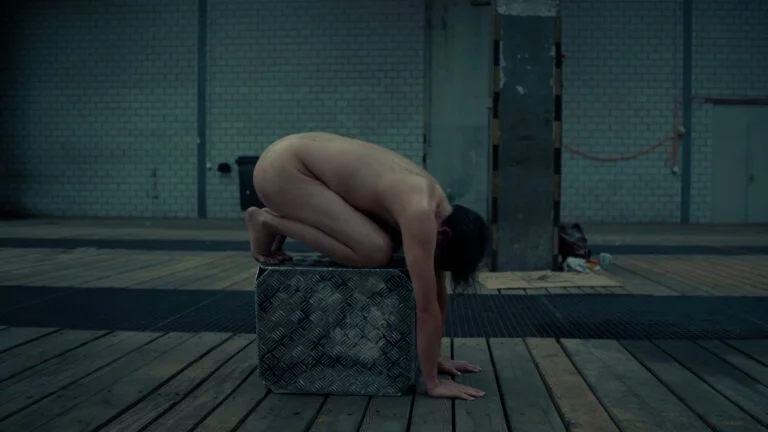 A nude performer is crouching on a textured metal block in an expansive wooden-floored space with brick walls and scattered objects in the background, embodying a contorted pose.