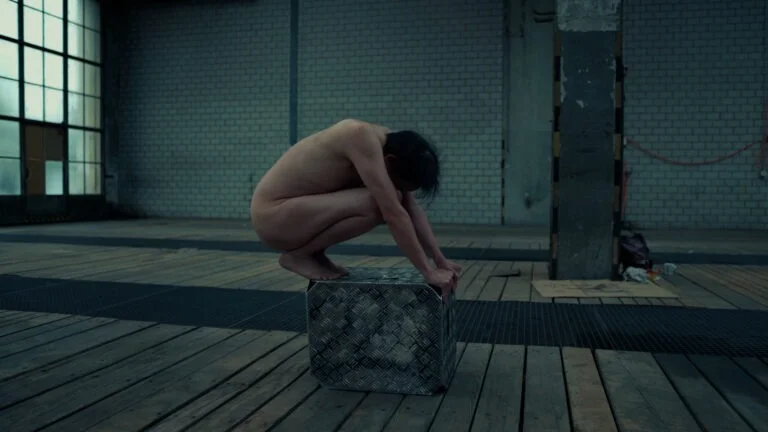 A nude performer squats on a metallic cube in an empty, dimly-lit industrial space with wooden flooring and large windows in the background, embodying the performance art piece titled 'Kist.'