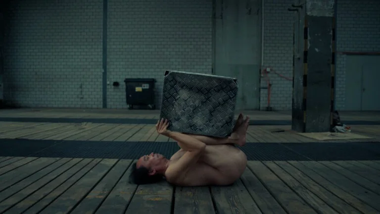 A nude performer balances a large metal box on their feet while lying on a wooden floor in an industrial space, with exposed brick walls and a dumpster in the background; this performance art piece is titled 'Kist.'