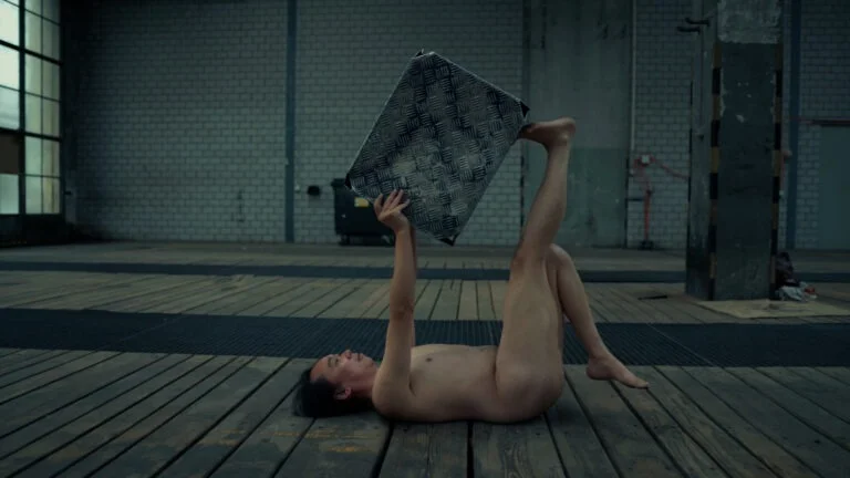 A naked performer lies on a wooden floor, holding a patterned metal sheet above them with one hand and both feet, in an industrial setting with large windows and concrete walls, as part of the performance art piece titled 'Kist'.