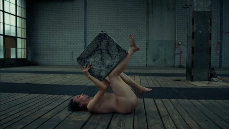 A performance art piece titled 'Kist' featuring a nude individual on a wooden floor, holding a large, textured metal object above their head while lying on their back. The setting has industrial elements, including large windows and exposed walls.