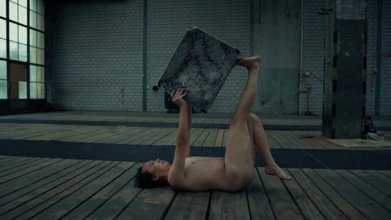 A naked performer lying on a wooden floor in an industrial space, holding a metallic patterned sheet above their head with both hands and feet. The environment is spacious with large windows and exposed brick walls, hinting at a contemporary art performance titled 'Kist.'