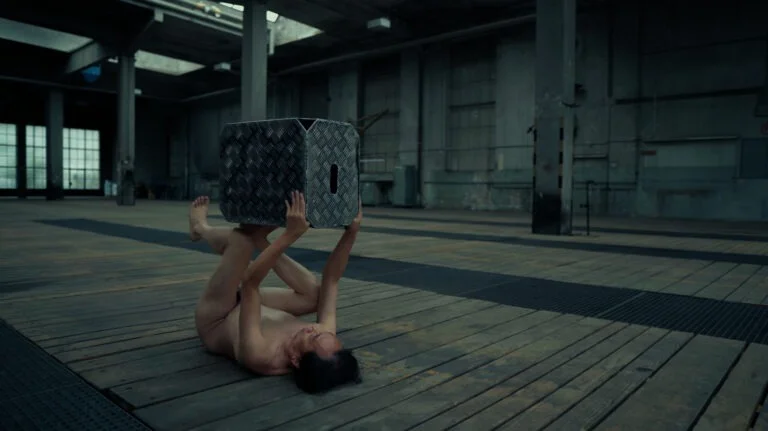 A naked performer engages in a contemporary art performance titled 'Kist,' lying on a wooden floor while holding a metallic box above their body in a large, industrial space.