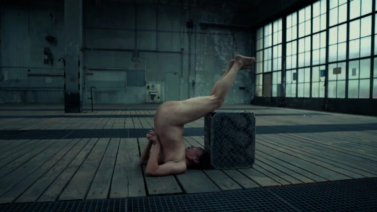 A naked performer titled 'Kist' is positioned upside down on a wooden floor, balancing on their head while their legs are raised and supporting a metallic box. The setting is an industrial space with large windows and a textured floor.