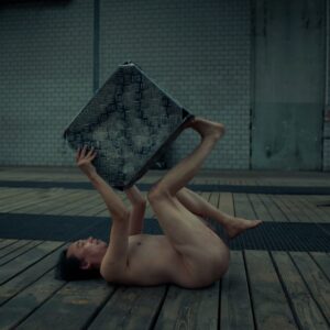 A nude performer lying on a wooden floor in an industrial space, holding a textured metal object above their head with both hands and their legs bent in the air. The backdrop features large windows and a brick wall.