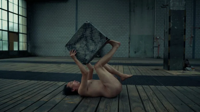 A nude performer lying on a wooden floor in an industrial space, holding a textured metal object above their head with both hands and their legs bent in the air. The backdrop features large windows and a brick wall.
