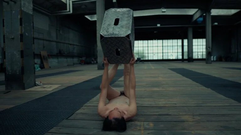 A performance artist named 'Kist' lies on a wooden floor, holding a large, metallic object with both hands above their head while remaining nude. The setting is an industrial space with large windows in the background and a minimalist aesthetic.