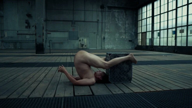 A nude performer balances their body upside down, with their head resting on the ground and legs bent backwards, positioned against a metallic box in a spacious industrial setting with wooden flooring and large windows.