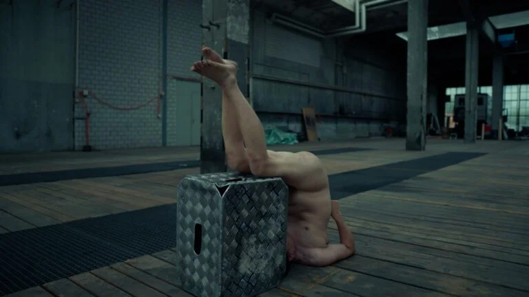 A naked performer, engaged in a performance art piece titled 'Kist', is balanced upside down on a metal box in an industrial space with wooden floors. The setting features concrete walls and scattered materials, enhancing the rawness of the performance.