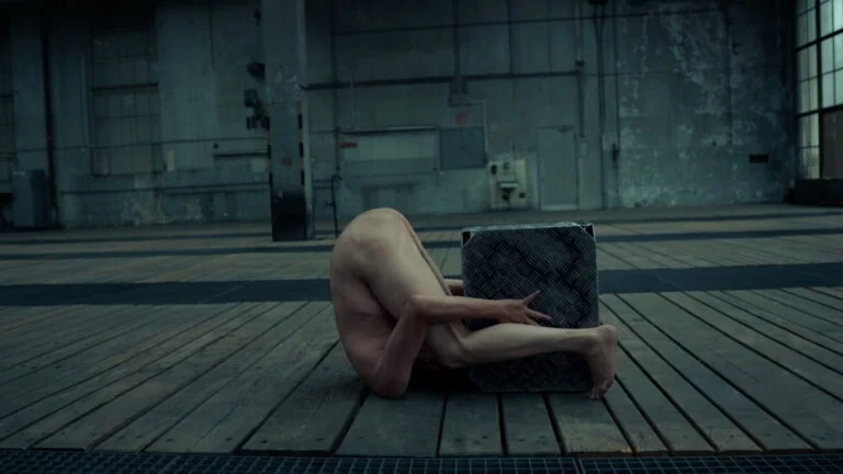 A naked performer, named Kist, contorts their body in a dynamic pose while partially enveloping a metal box in an industrial space with wooden flooring and gray walls.