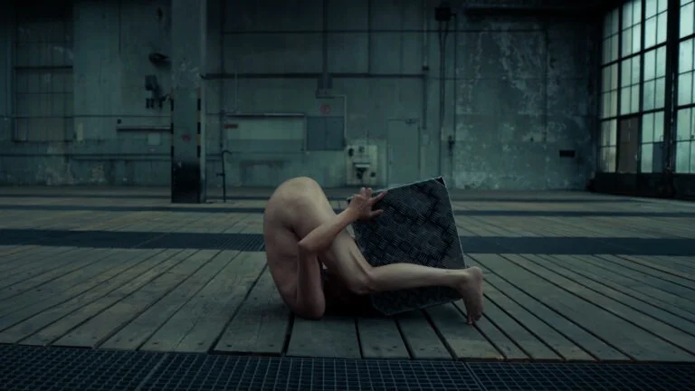 A nude performer in a warehouse space contorts their body around a metal square, creating a striking pose against the wooden floor and concrete walls, representing the performance art piece 'Kist.'