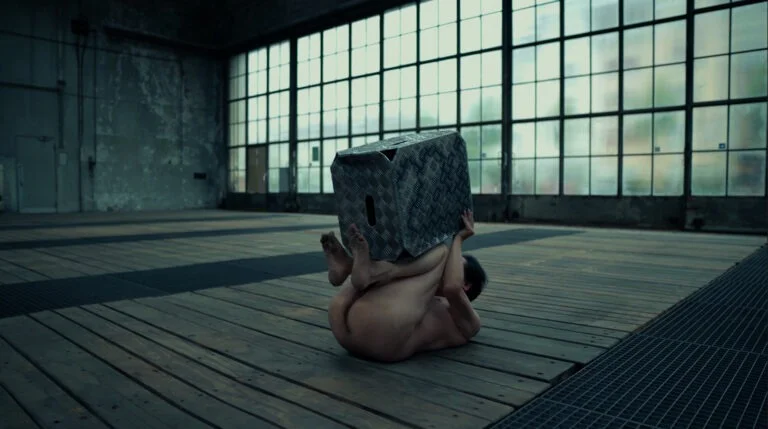 A naked performer is curled up on a wooden floor, balancing a textured metal box on their head, set in an industrial space with large windows.