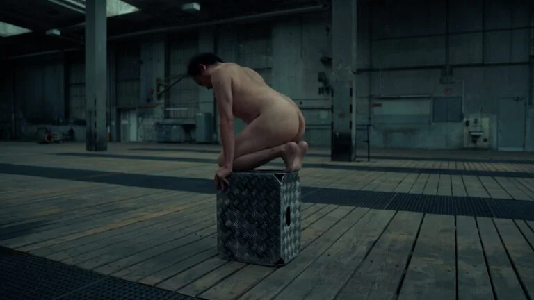 A naked performer is crouching on a metallic stool in an industrial space with wooden flooring, surrounded by a sparse, empty setting that features large windows and concrete walls, representing the performance art piece titled 'Kist.'