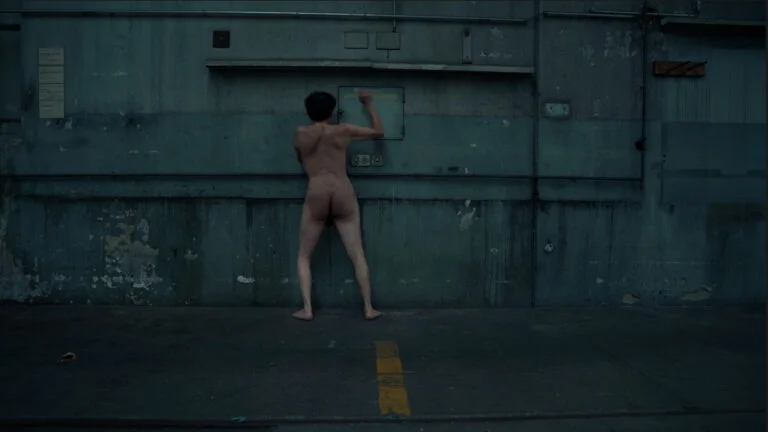 A nude performer named Kist stands against a weathered blue-green wall, engaging with a blank panel while positioned in profile, with their back turned to the viewer. The industrial setting features peeling paint and visible electrical outlets, adding to the stark atmosphere.