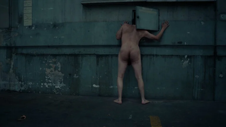A naked performer leans into a small opening in a concrete wall, their head obscured, in a performance art piece titled 'Kist.' The setting features a weathered, industrial background with peeling paint and a muted color palette.