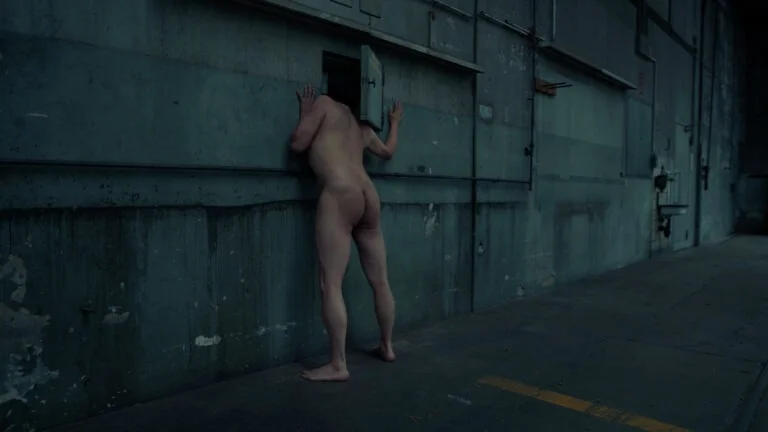 A naked individual leans against a textured wall, peering through a small opening, in a dimly lit, industrial setting, as part of the performance art piece titled 'Kist.'
