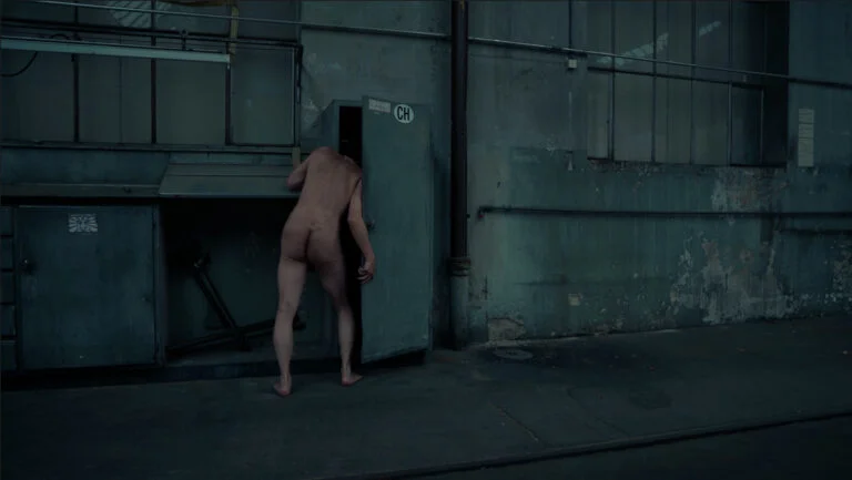 A naked individual is bent over, partially entering a grey metal cabinet in an industrial setting, with peeling paint and exposed pipes on the walls, titled 'Kist'.