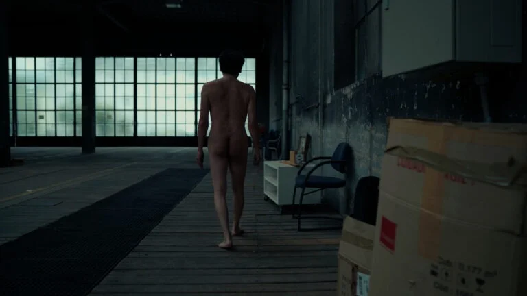 A naked performer titled 'Kist' walks away from the camera in an industrial space, surrounded by boxes and furniture, with large windows in the background providing a muted light.