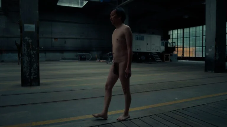 A naked performer named Kist walks barefoot in a dimly lit industrial space, characterized by concrete walls and large windows, with a minimalistic interior featuring a few scattered objects.