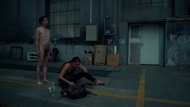 A naked performance art scene titled 'Kist,' featuring a man standing to the left and a woman sitting on the ground to the right, focused on a laptop and surrounded by scattered objects. The industrial background includes large windows and exposed walls, enhancing the raw atmosphere.
