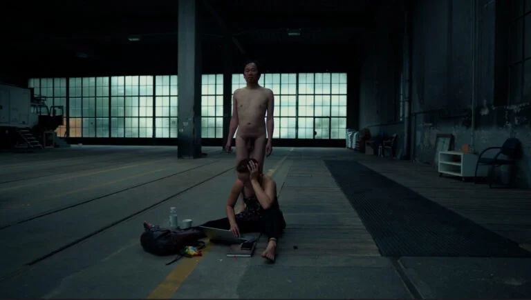 A performance art piece titled 'Kist,' featuring a naked person standing on stage while another individual sits on the floor, facing downward with their hands on their head, surrounded by personal items including a backpack and a laptop. The setting is a spacious, industrial-looking room with large windows in the background.