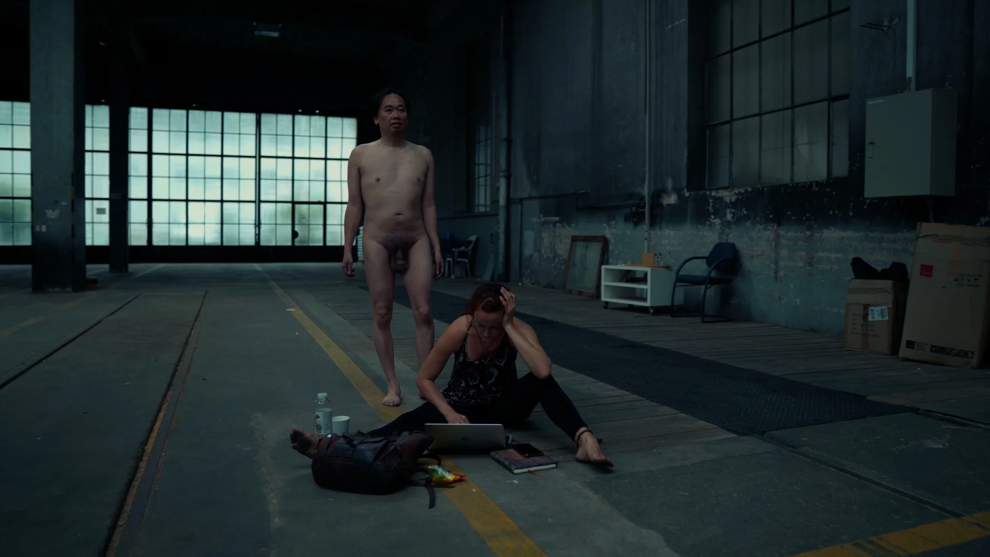 A performance art piece titled 'Kist' features a naked male figure standing in the background, while a seated female figure appears distressed while working on a laptop. The setting is an industrial space with large windows and minimal furnishings, including a backpack and scattered items on the floor.