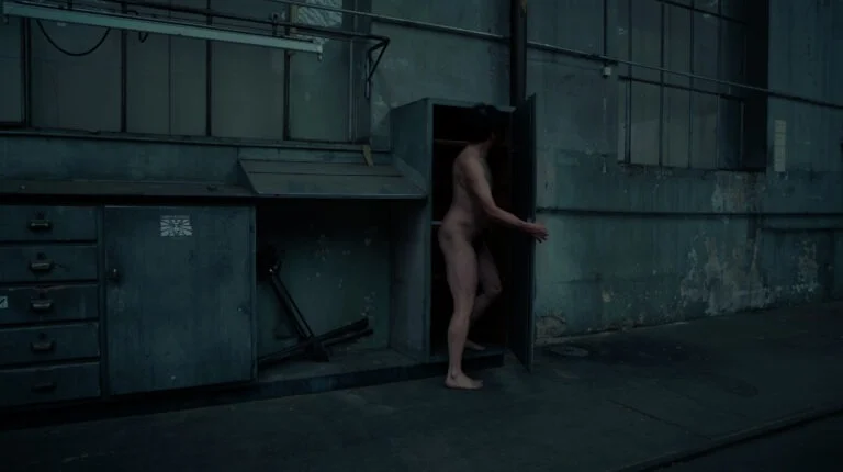 A naked performer, part of an artwork titled 'Kist', emerges from an open metal cabinet in an industrial setting with peeling green walls and large windows. The figure is shown in profile, displaying a subtle interaction with the surrounding space.