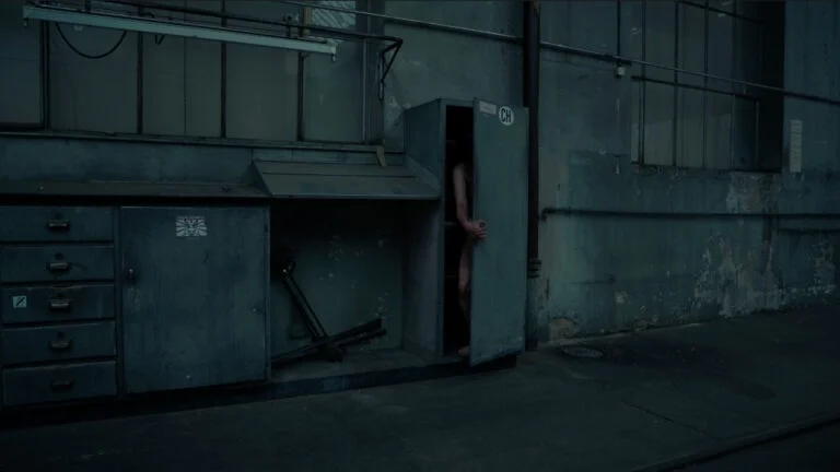A naked performer partially emerges from a metal cabinet in an industrial setting, showcasing a contrast between the human form and the surrounding machinery and walls, which are faded and worn.